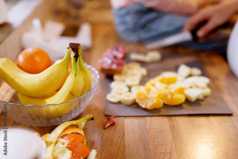 Close up focus view of orange and bananas for fruit salad and little toddler girl sitting on the kitchen table and waiting while her pregnant mum cutting an apple.