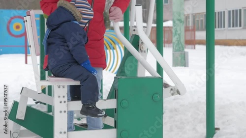 A cute little boy and a young mother are engaged in outdoor exercise equipment. It's hard for a kid to reach the handles of equipment