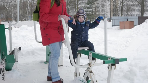 A cute little boy and a young mother are engaged in outdoor exercise equipment. It's hard for a kid to reach the handles of equipment