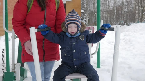 A cute little boy and a young mother are engaged in outdoor exercise equipment. It's hard for a kid to reach the handles of equipment