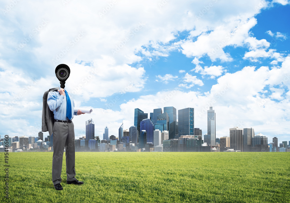 Camera headed man standing on green grass against modern cityscape Camera headed man standing on green grass against modern cityscape
