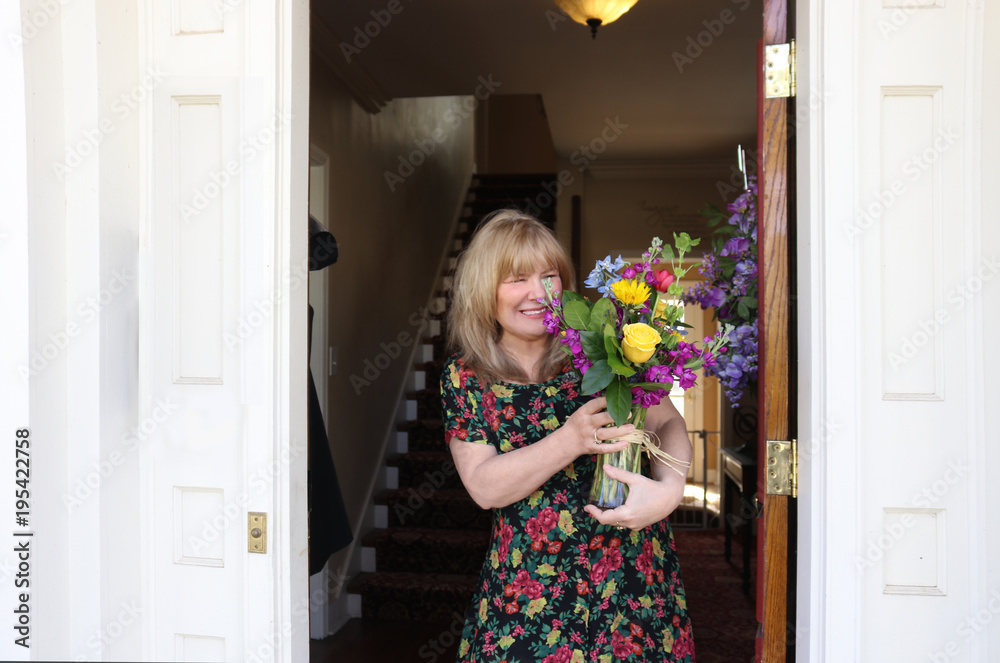 Pretty blond middle ages woman in flowered dress looks delighted with flowers that have just been delivered - standing in door of house