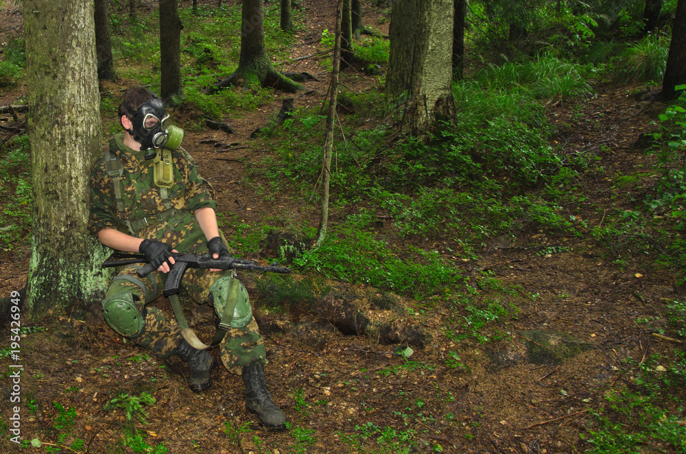 soldier with a rifle and a gas mask sits on a fallen forest tree soldier with a rifle and a gas mask sits on a fallen forest tree