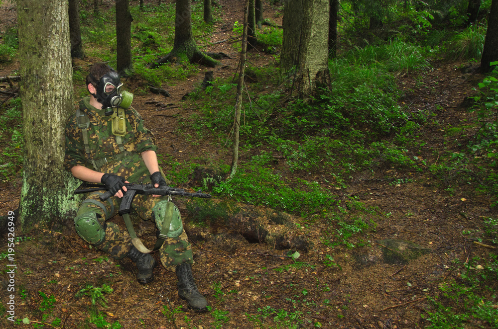 soldier with a rifle and a gas mask sits on a fallen forest tree