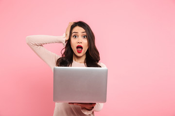 Image of thrilled woman 20s in casual clothing shouting and grabbing head while using silver notebook, isolated over pink background