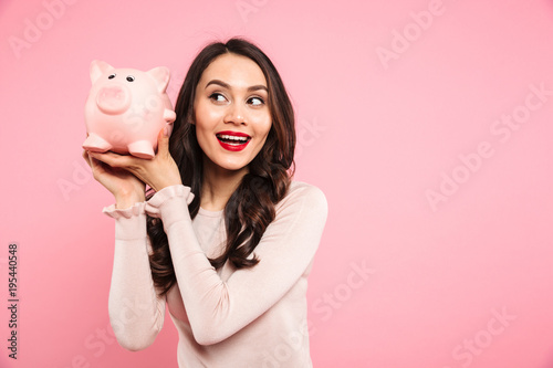 Fotografia Joyous young woman 20s in casual clothing holding piggybank with lots of money,
