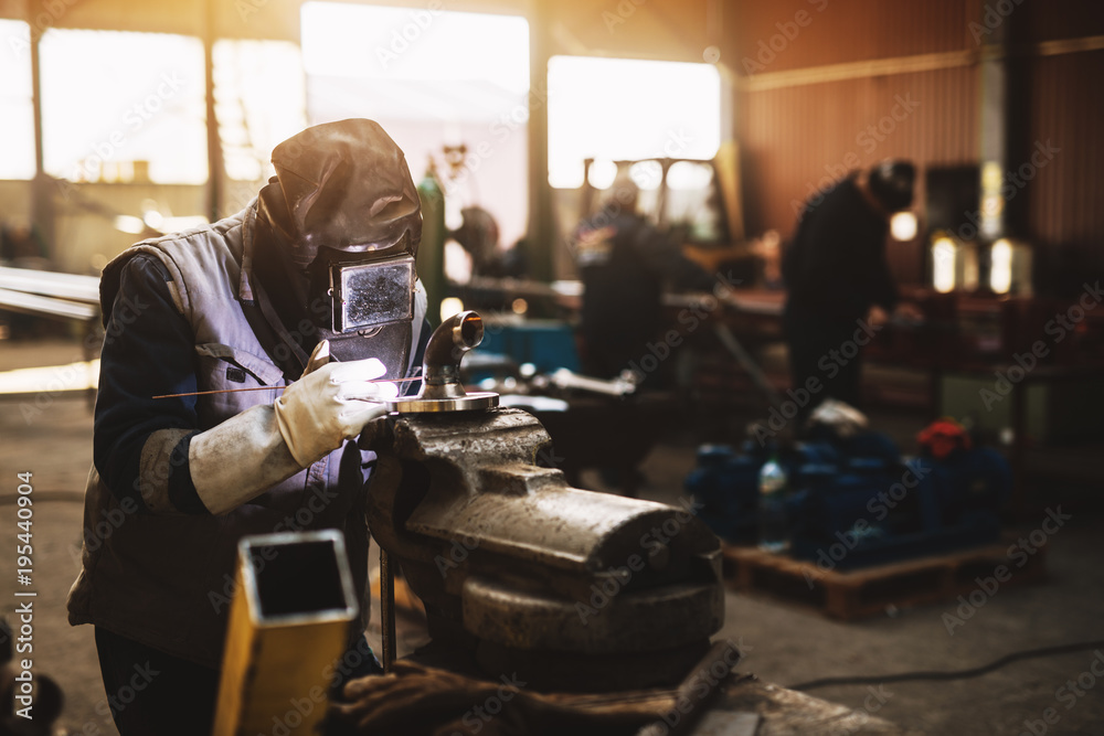Welder in protective uniform and mask welding metal pipe on the industrial table while sparks flying. Welder in protective uniform and mask welding metal pipe on the industrial table while sparks flying.