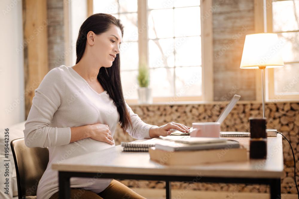 Difficult work. Charming serious pregnant woman sitting in profile while using laptop and putting hand on belly Difficult work. Charming serious pregnant woman sitting in profile while using laptop and putting hand on belly