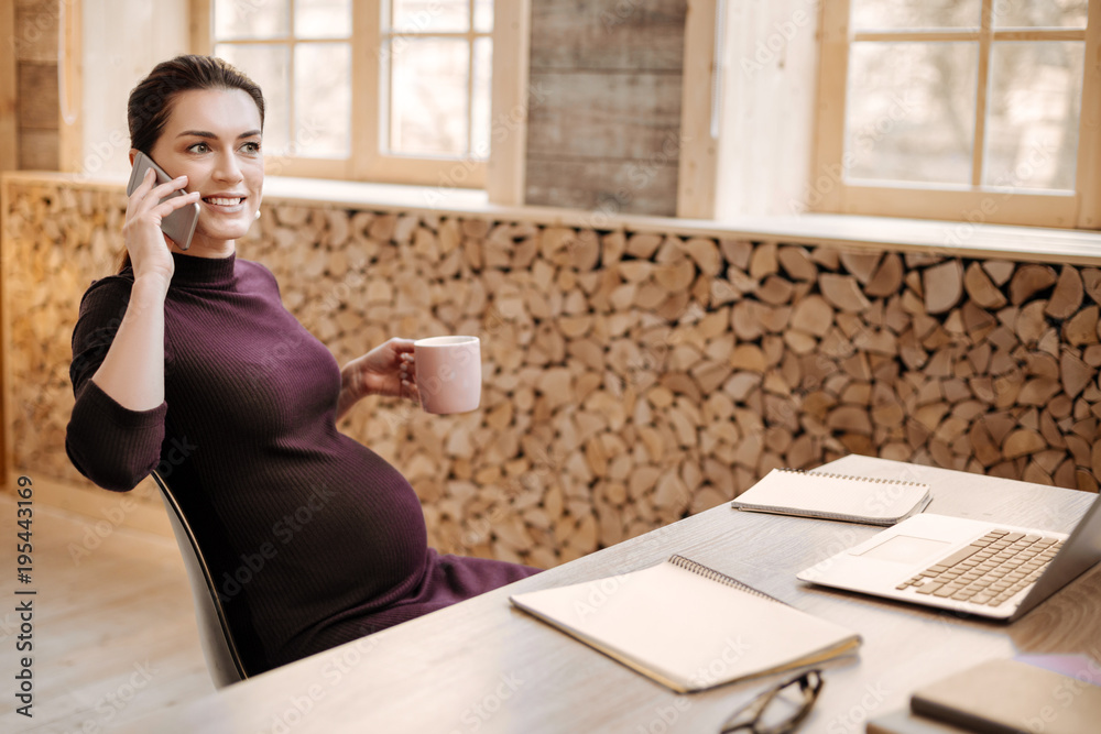 Business negotiation. Appealing positive pregnant businesswoman making call while holding mug and posing at table Business negotiation. Appealing positive pregnant businesswoman making call while holding mug and posing at table