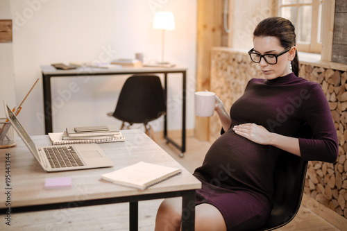 Baby in belly. Pretty attractive pregnant businesswoman caressing belly while elevating mug and talking to baby