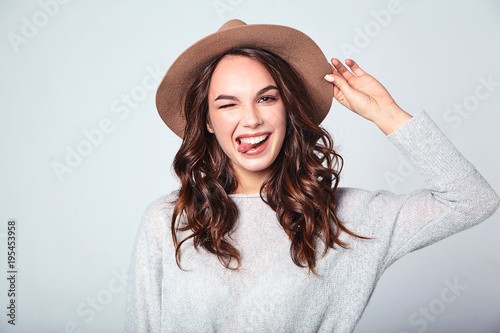 Portrait of young stylish laughing girl model in gray casual summer clothes in brown hat with natural makeup isolated on gray background. Looking at camera and showing her tongue