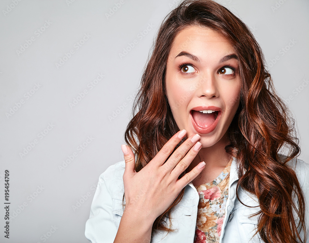 Portrait of young stylish laughing girl model in colorful casual summer clothes with natural makeup isolated on gray background. Looking at camera with hands near mouth Portrait of young stylish laughing girl model in colorful casual summer clothes with natural makeup isolated on gray background. Looking at camera with hands near mouth