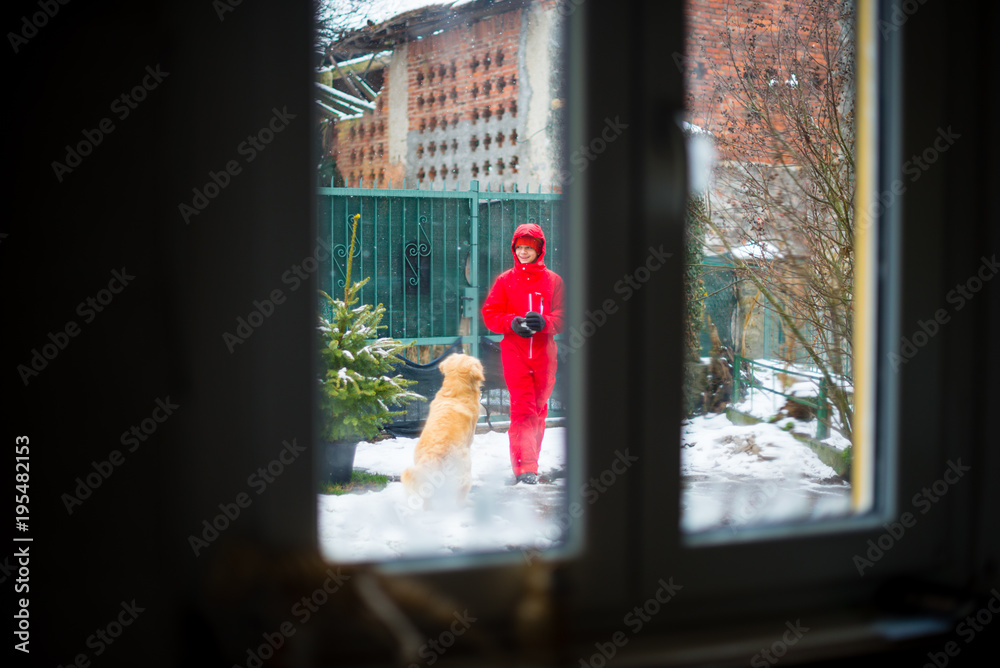 boy plays in the garden with the snow and with his dog, seen from the window inside the house