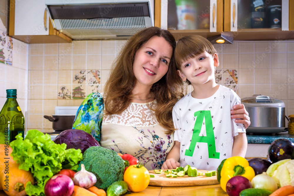 The young cook mother standing with her little son in the kitchen and salting vegetables