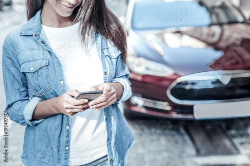Modern device. Nice attractive pleasant woman standing in front of her car and holding her smartphone while using it