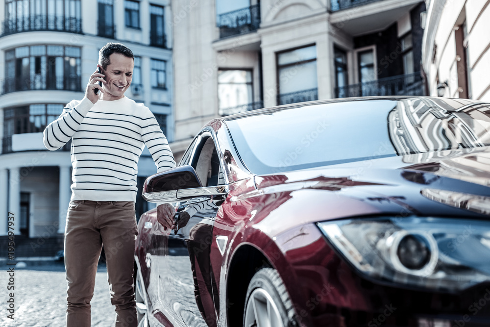 Fototapeta premium Ready to go. Delighted joyful nice man standing near his automobile and talking on the phone while being ready to go