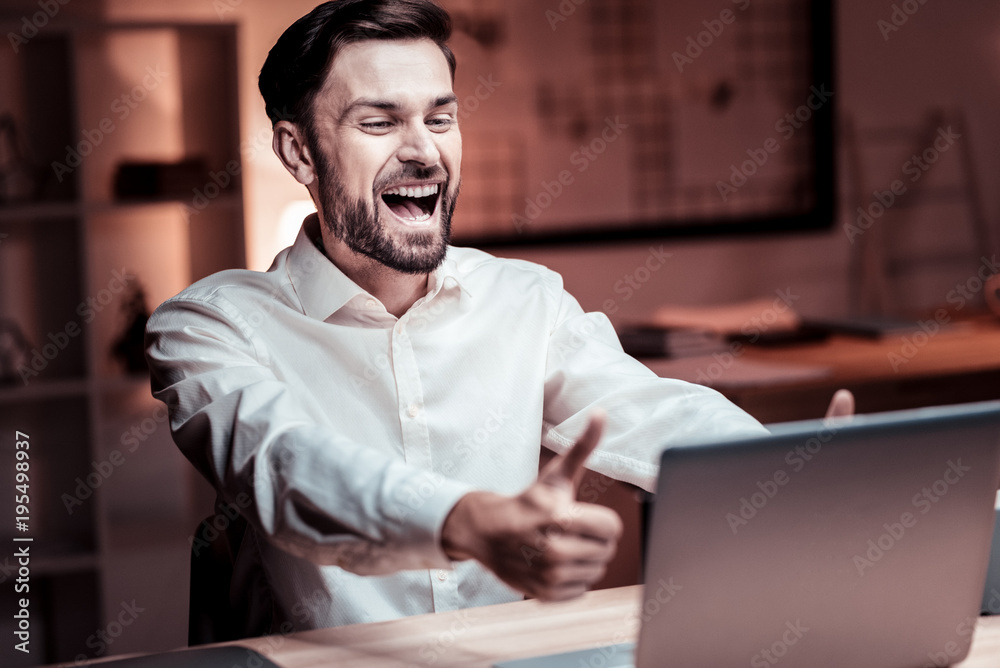 Well done. Satisfied occupied unshaken man using laptop in his cabinet rejoicing and laughing. Well done. Satisfied occupied unshaken man using laptop in his cabinet rejoicing and laughing.
