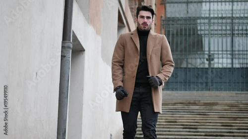 Handsome bearded young man outdoor in winter fashion, walking up marble stairs wearing black turtleneck sweater and woolen blazer jacket in city setting