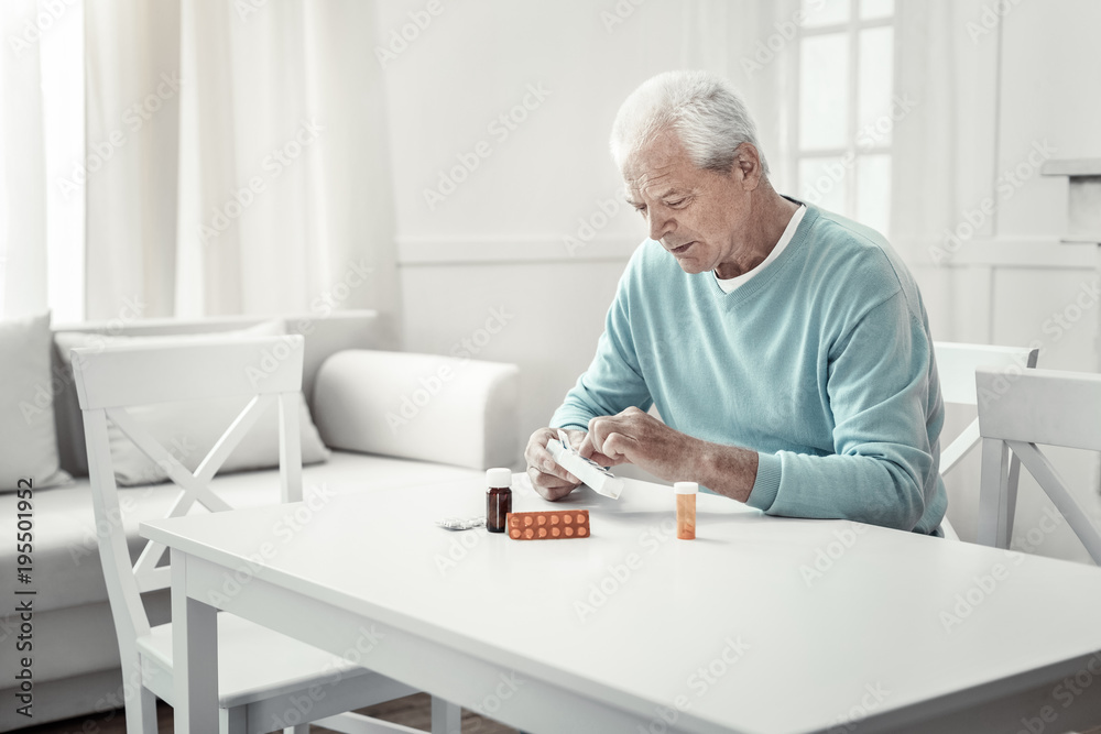 Struggle with the illness. Lonely serious aged man sitting in the bright room holding pack of tablets and overlooking pills. Struggle with the illness. Lonely serious aged man sitting in the bright room holding pack of tablets and overlooking pills.