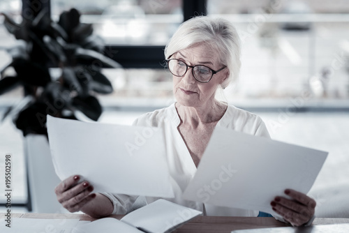 Need to think. Attentive female person holding papers in her hands and raising eyebrows while reading information