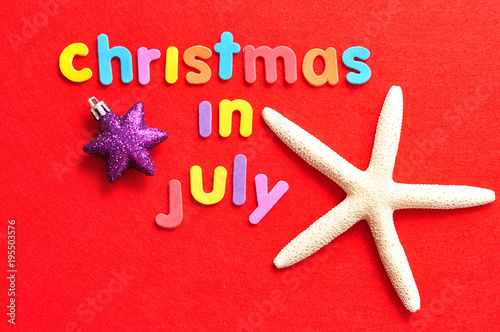 The words christmas in July in colorful letters on a red background with a starfish and a star shape bauble