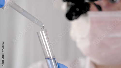 Laboratory work  a woman doctor holds a test tube  in a medical mask and professional glasses  a magnifying glass  takes an analysis from a test tube in blue rubber gloves  analysis  blood  DNA  a tes