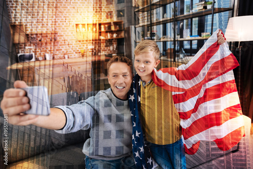 Proud Americans. Emotional young man smiling while sitting near his happy son with a flag of the USA and feeling glad while taking photos after coming to the country
