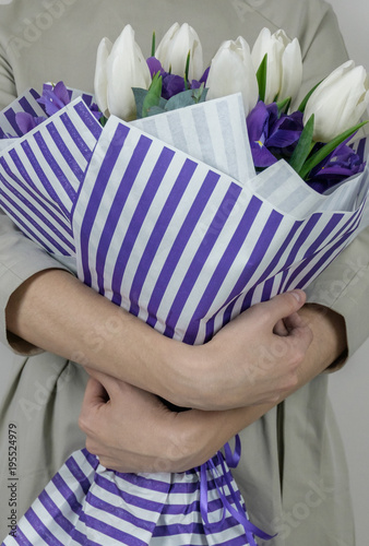 Unrecognizible woman in beige dress holding bouquet of white tulips and violet iris in striped lilac wrapping paper.