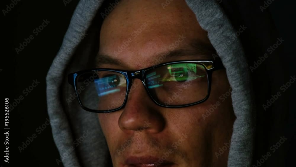 Hacker in glasses and a hood works behind a computer in the dark, the program code is reflected in glasses close-up, 4k Hacker in glasses and a hood works behind a computer in the dark, the program code is reflected in glasses close-up, 4k