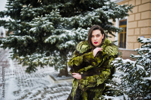 Brunette girl in green fur coat at winter day against snowy pine tree.