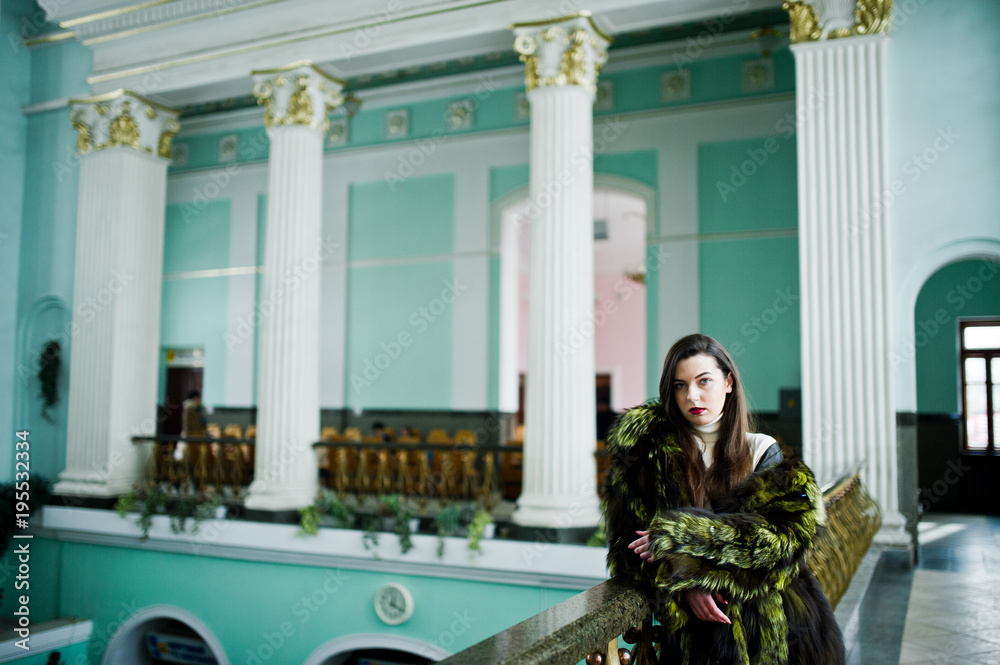 Brunette girl in green fur coat in old hall with column and railings.