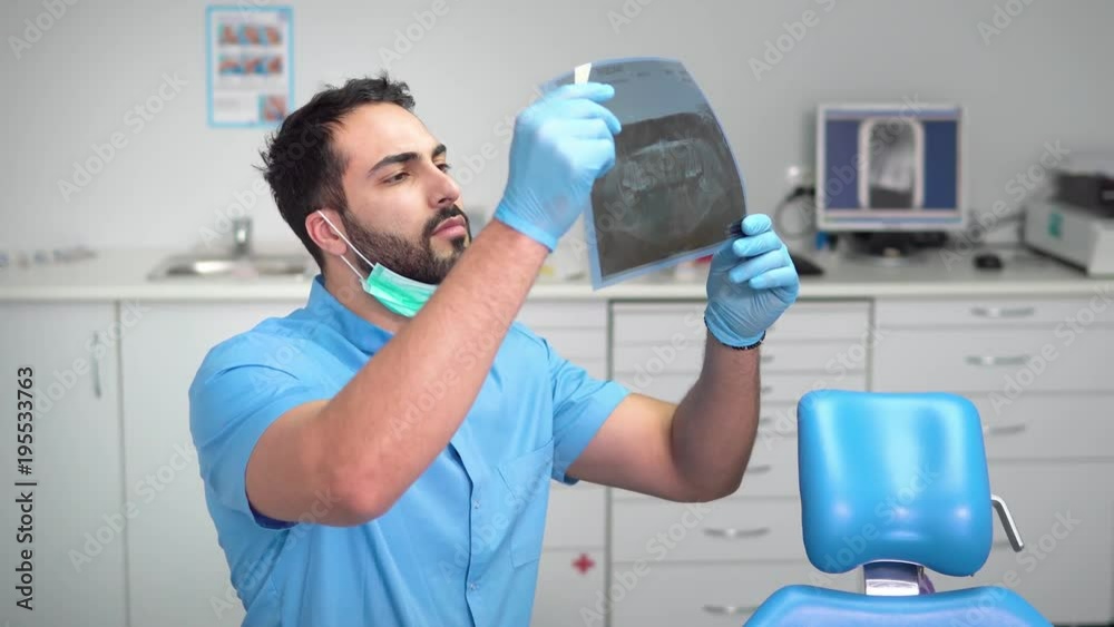 Intelligent dentist stydying an x-ray at the workplace, black-haired man in blue uniform looking attentively at the photograph, sitting near dentist chair in well-equipped office Intelligent dentist stydying an x-ray at the workplace, black-haired man in blue uniform looking attentively at the photograph, sitting near dentist chair in well-equipped office