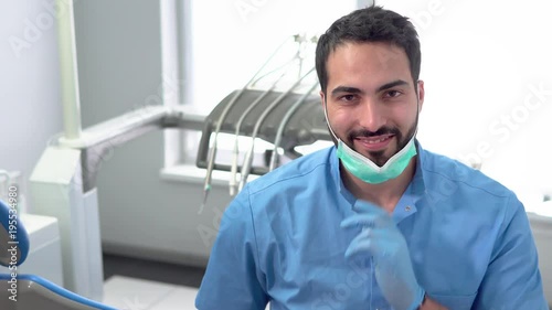 Portrait of professional dentist in the office, black-haired man in blue gloves holding a false teeth sample, smiling doctor taking off the mask