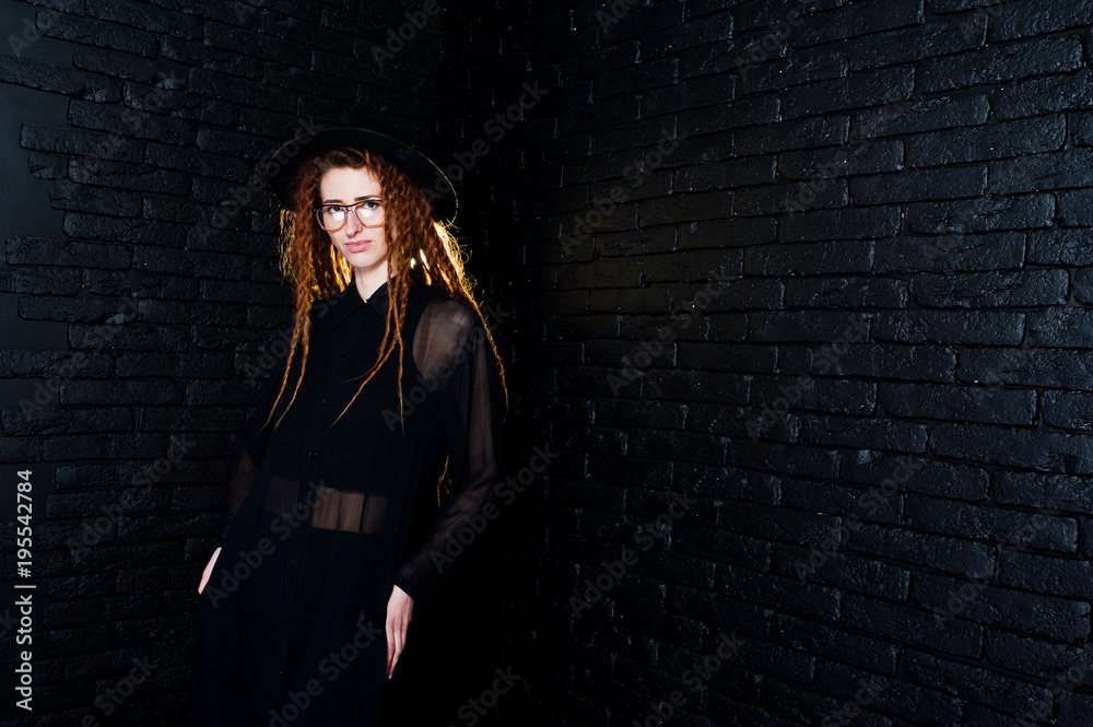 Studio shoot of girl in black with dreads, at glasses and hat on brick background.