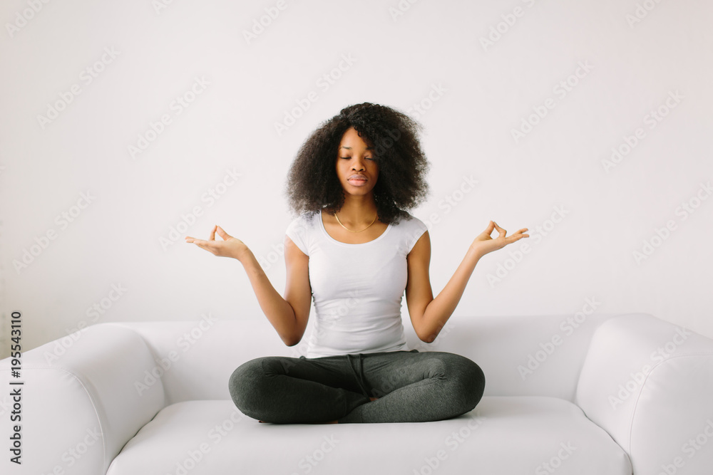 An African American young women sitting in the lotus position on white bed An African American young women sitting in the lotus position on white bed