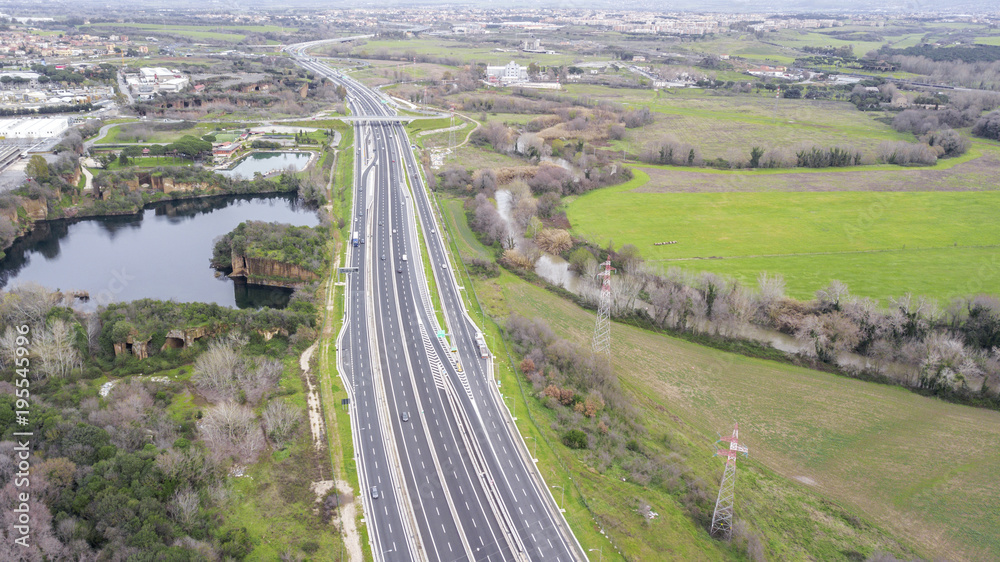 Aerial view of a stretch of highway in the Italian countryside. The road is divided into many lanes in each direction. There are cars and trucks on the street.Around the road there are trees and grass Aerial view of a stretch of highway in the Italian countryside. The road is divided into many lanes in each direction. There are cars and trucks on the street.Around the road there are trees and grass