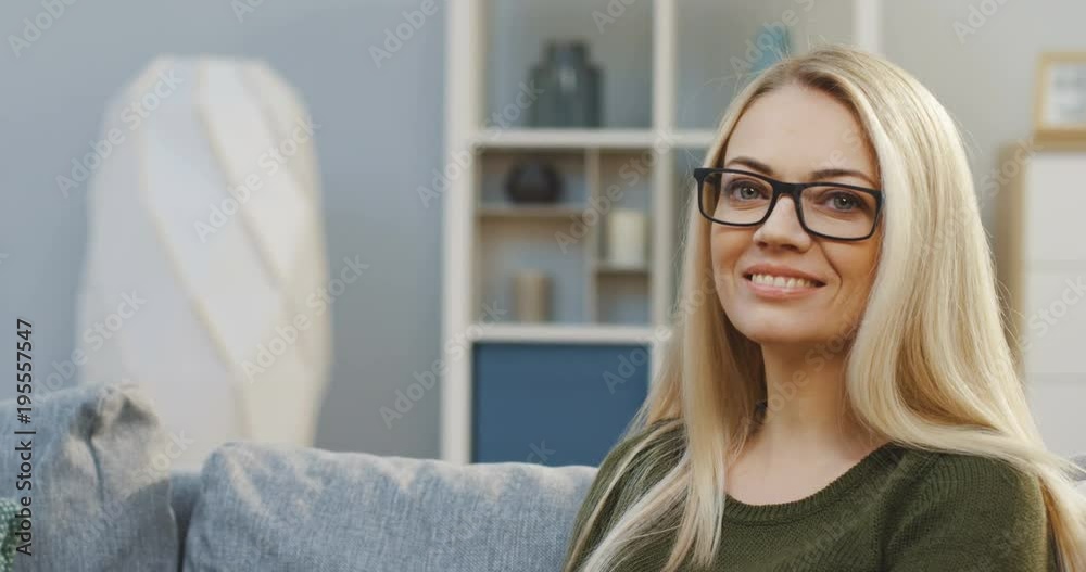 Portrait shot of the blonde charming woman in glasses turning her head to the camera and smiling in the living room. Close up. Inside