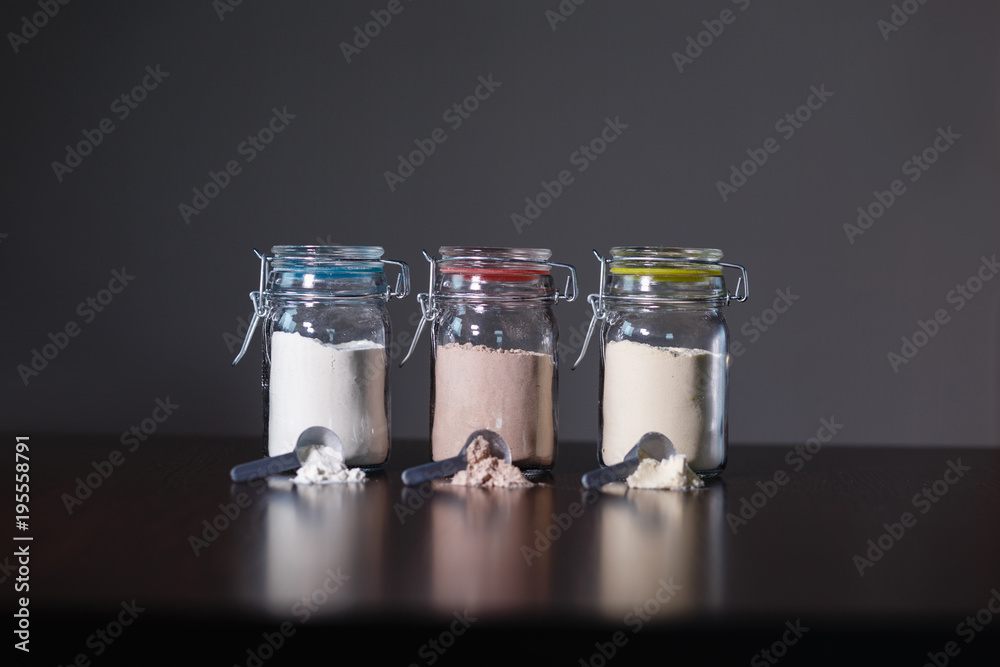 Three glass jars with protein powder, strawberries, banana and chocolate and three scoops for milkshakes. Sports and dietary nutrition for athletes and weight loss or muscle mass bodybuilders. Three glass jars with protein powder, strawberries, banana and chocolate and three scoops for milkshakes. Sports and dietary nutrition for athletes and weight loss or muscle mass bodybuilders.