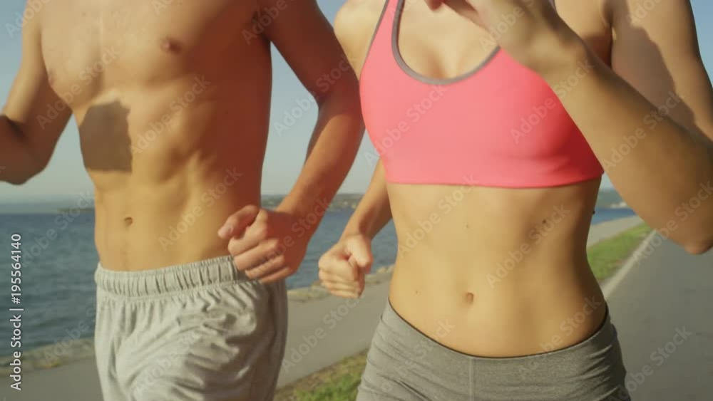 SLOW MOTION, CLOSE UP: Shirtless man and woman wearing sports clothing run along sunlit summer coastline. Slow motion shot of athletic couple's bodies moving during their jog down empty ocean road. SLOW MOTION, CLOSE UP: Shirtless man and woman wearing sports clothing run along sunlit summer coastline. Slow motion shot of athletic couple's bodies moving during their jog down empty ocean road.