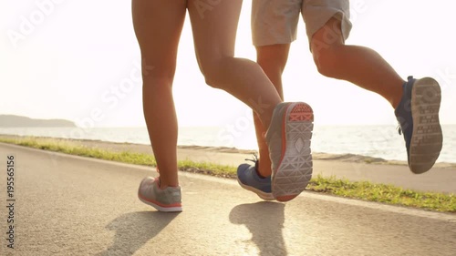 SLOW MOTION, LOW ANGLE, SUN FLARE: Young athletic woman and man wearing training shoes run along sunny ocean road. Unrecognizable Caucasian girlfriend and boyfriend jog on a sunny day at the beach.