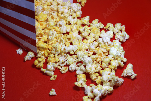 Heap of popcorn from red and white paper box on the red floor. It is the corn of a variety with hard kernels that swell up and burst open with a pop when heated.