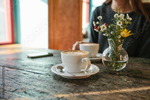Obraz na płótnie Two cups of coffee and a vase of flowers on a wooden table, the girl holds in her hand one cup of coffee in the background. A photo indicates a meeting of people and a joint pastime. w salonie