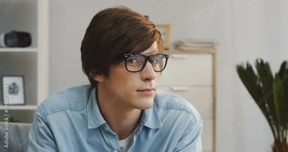 Close up of the attractive young man in glasses turning his head and smiling sincerely to the camera in the modern room of the apartment. Inside. Portrait Close up of the attractive young man in glasses turning his head and smiling sincerely to the camera in the modern room of the apartment. Inside. Portrait