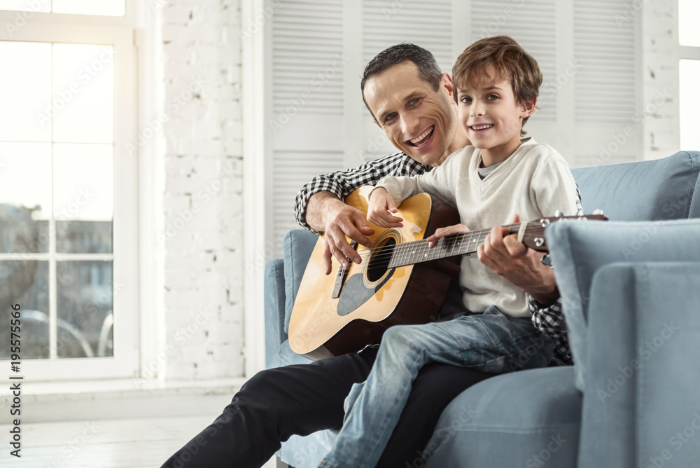 Great day. Nice alert fair-haired boy learning to play the guitar while sitting on the couch and his father smiling Great day. Nice alert fair-haired boy learning to play the guitar while sitting on the couch and his father smiling