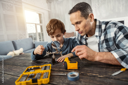 Constructing. Good-looking concentrated loving dark-haired man showing instruments to his son while sitting at the table and hisson looking at a magnifying glass