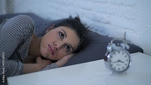 lateral panning shot of young attractive hispanic woman upset in stress and insomnia lying anxious on bed trying to sleep suffering sleeping disorder at night looking alarm clock frustrated 