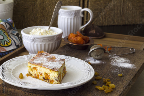 Cottage cheese casserole with raisins and dried apricots. A piece of casserole with powdered sugar on a white plate, fresh farmer cottage cheese in a white bowl on a wooden table. Rustic style.