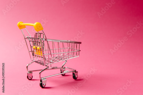 Close up of supermarket grocery push cart for shopping with black wheels and yellow plastic elements on handle isolated on pink background. Concept of shopping. Copy space for advertisement