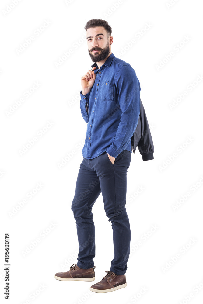 Side view of young bearded man in blue shirt carrying bomber jacket over the shoulder looking at camera. Fully body isolated on white background. Side view of young bearded man in blue shirt carrying bomber jacket over the shoulder looking at camera. Fully body isolated on white background.