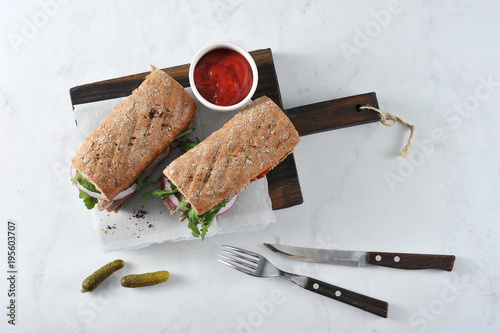 Sandwich on a wooden board. The filling of the sandwich consists of beef, arugula, red onion and marinated cucumber slices. In the frame, cucumbers, a cup of ketchup, cutlery. View from above.